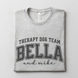 May include: A light gray t-shirt with the text "THERAPY DOG TEAM" above the name "BELLA" in bold, block letters, and "and mike" in a script font. The shirt has a crew neck and is folded.