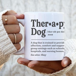 May include: White ceramic mug with the words "Therapy Dog" in a bold, black font. The mug includes a definition of a therapy dog. The mug is held by a person wearing a cream-colored sweater.
