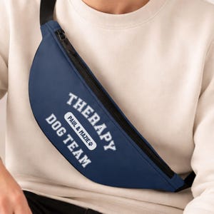May include: Navy blue fanny pack with a black zipper and strap. The text "THERAPY DOG TEAM" is printed in white, with "PAUL & MAZIE" in a smaller oval. A practical accessory for dog lovers.