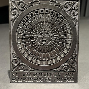 Peut inclure: Un cadran solaire rectangulaire de couleur argentée avec une bordure gravée ornée. Le cadran central représente un visage de soleil et un anneau de chiffres représentant les jours du mois. Des chiffres romains sont gravés en bas.