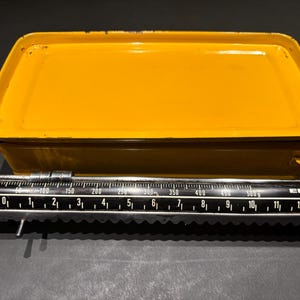 Puede incluir: Una báscula de cocina amarilla vintage con una barra de medición negra y plateada. La báscula tiene una bandeja y un rango de medición de hasta 12 kg. La marca "EKS" es visible.