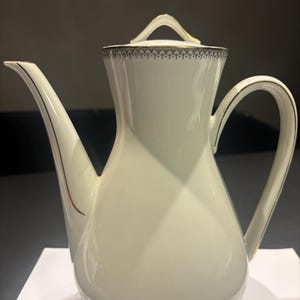 Può includere: Una caffettiera in porcellana bianca con beccuccio e manico curvi. La caffettiera ha un bordo dorato lungo il bordo, il beccuccio e il manico. Un motivo decorativo nero e oro circonda la parte superiore della caffettiera.