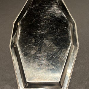 Può includere: Un vassoio ottagonale color argento con un bordo leggermente rialzato. Il vassoio ha una superficie riflettente con graffi visibili, che suggeriscono che sia vintage o ben usato. La forma del vassoio è geometrica, con lati dritti e angoli angolati.