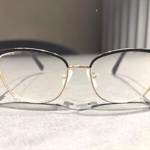 May include: Cat-eye eyeglasses with a black upper frame and gold-colored lower frame. The lenses are clear, and the glasses are set against a neutral background. The frames have a delicate, modern design.
