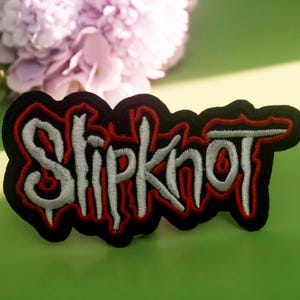 Pode incluir: Patch bordado com o nome da banda "Slipknot" em branco, contornado em vermelho, sobre um fundo preto. O patch tem uma borda irregular e está sobre uma superfície verde com uma flor e um pirulito.