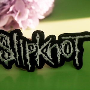May include: A black embroidered patch with the word "Slipknot" in white, stylized lettering. The patch is on a green surface, with a blurred purple flower in the background. A colorful candy is in the background.