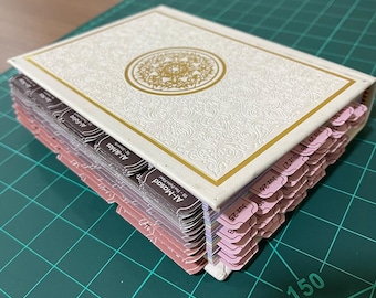 May include: A cream-colored book with gold accents and a decorative embossed cover. The book has labeled tabs in shades of pink and brown. The tabs are labeled with text. The book is open, revealing the pages.