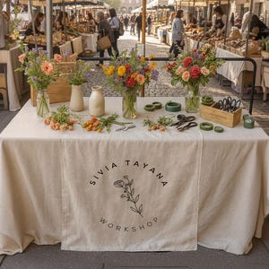May include: A flower market stall displays colorful bouquets in glass vases, along with floral arranging tools and supplies. The table is covered with a cream-colored cloth, featuring the text "SIVIA TAYANA WORKSHOP" in a circular design.