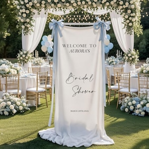 May include: A white banner with the text "WELCOME TO AURORA'S Bridal Shower MARCH 28TH, 2026" is displayed at an outdoor event. The banner is tied with light blue ribbons. The event features floral arrangements, tables, chairs, and a decorative arch.