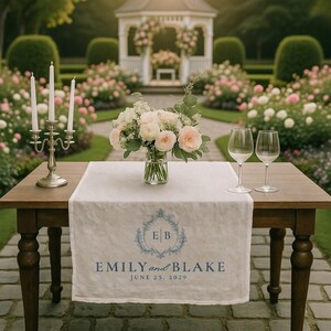 May include: A wooden table set for a wedding ceremony, with a white linen runner featuring the names "EMILY and BLAKE" and the date "JUNE 25, 2029". A floral arrangement and two wine glasses are on the table, with a gazebo and rose garden in the background.
