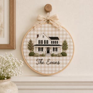 May include: A circular embroidery hoop featuring a beige and white gingham fabric background. Centered is a detailed stitched illustration of a two-storey house with black shutters and a porch, with the embroidered text "The Evans" below it.