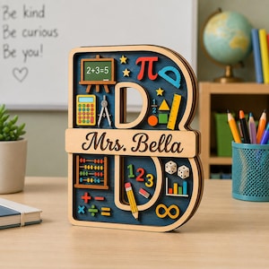 May include: A wooden letter B sign featuring a dark blue background with colorful educational icons including a chalkboard with 2+3=5, a calculator, abacus, dice, math symbols, and a pencil. A horizontal wooden bar across the center displays the text Mrs. Bella.