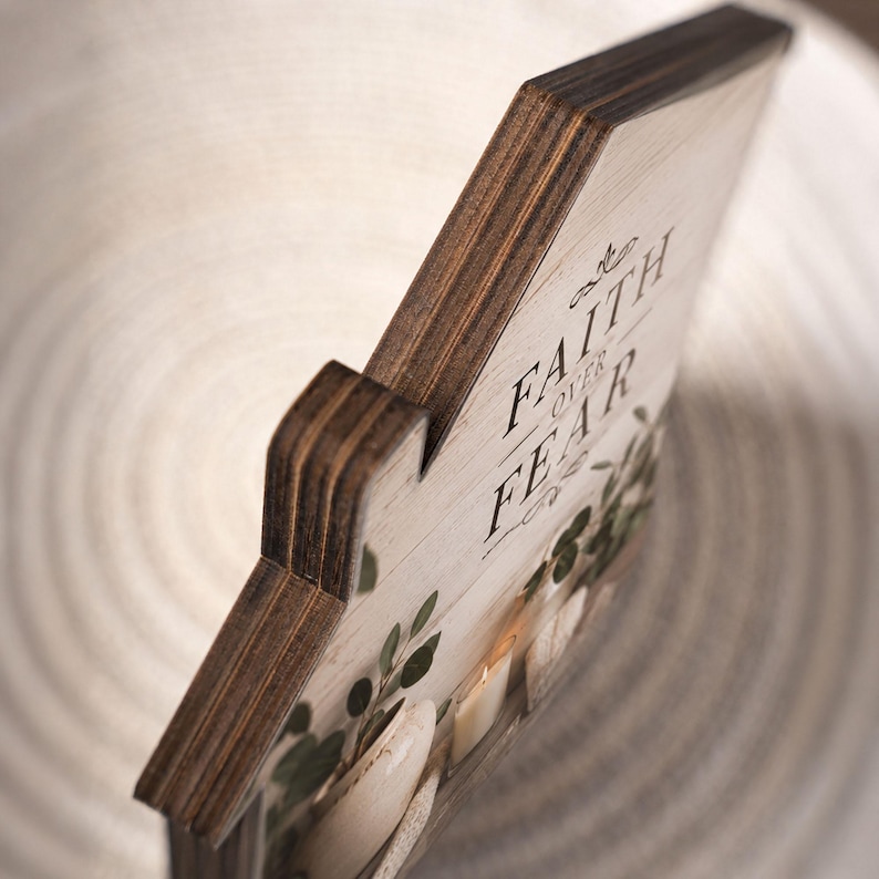 Puede incluir: Un letrero de madera con forma de casa, bordes te&ntilde;idos oscuros y frente de veta clara. El texto "FAITH OVER FEAR" est&aacute; impreso en negro, acompa&ntilde;ado por un gr&aacute;fico de hojas verdes y piedras blancas en la parte inferior. Construcci&oacute;n de madera contrachapada.