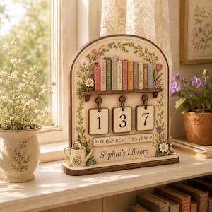 May include: A wooden arched tabletop tracker featuring a shelf with miniature book spines, three hanging number tiles displaying 137, and the text "BOOKS READ THIS YEAR" and "Sophia's Library". The frame is decorated with painted floral motifs.