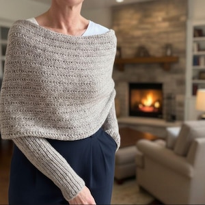 Puede incluir: Un chal de punto beige con mangas. El chal está hecho de un hilo grueso y texturizado y tiene un estilo amplio de cuello barco. Las mangas son largas y ajustadas. El chal se usa sobre una camiseta blanca y pantalones azul oscuro.