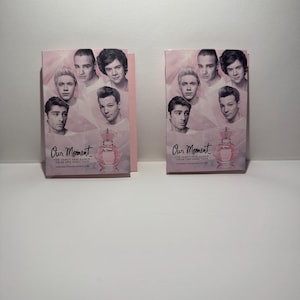 Puede incluir: Dos cajas de perfume rosas con el texto "Our Moment" y la frase "THE DEBUT FRAGRANCE FROM ONE DIRECTION". Las cajas presentan imágenes de los miembros de la banda y un frasco de perfume.