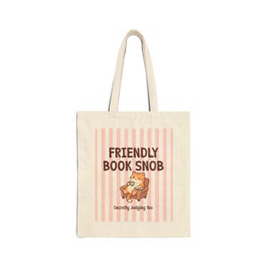 May include: Beige tote bag with a design featuring the text "FRIENDLY BOOK SNOB" above an illustration of a cat in a chair. The cat is holding a mug. Below the illustration is the text "Secretly Judging You".