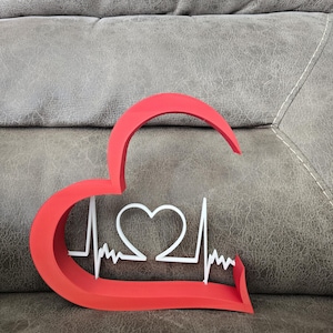 Op de afbeelding: Een rood hartvormig decoratief stuk met een wit hart en een hartslaglijn ontwerp. Het stuk is gemaakt van een solide materiaal en is geplaatst op een grijze ondergrond. Het ontwerp is een symbool van gezondheid en welzijn.