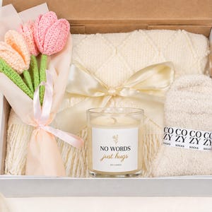 May include: A gift box containing a cream-colored diamond-patterned throw blanket tied with a satin ribbon, a pair of fuzzy cream socks labeled COZY SOCKS, a small glass candle with a label reading NO WORDS just hugs, and crocheted tulips.