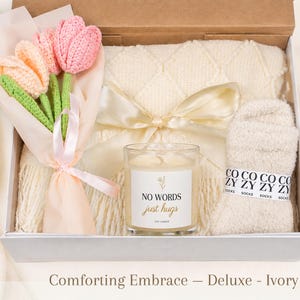 May include: A gift box containing a bouquet of three crocheted tulips in pink and peach, a cream-colored knit blanket tied with a satin ribbon, a glass candle with a label reading NO WORDS just hugs, and a pair of fuzzy cream-colored socks.