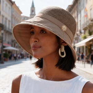 May include: A taupe sun hat with a textured crown and a wide, smooth brim. The hat features a matching fabric band around the base of the crown, accented with a small, circular, gold-toned metallic embellishment on the side of the hat.