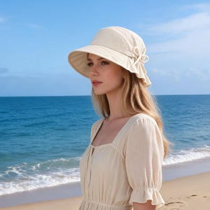 Könnte beinhalten: Ein beiger Sonnenhut mit breiter Krempe und dekorativer Schleife an der Seite. Die Person trägt ein beiges Kleid und steht an einem Sandstrand mit dem Meer im Hintergrund.
