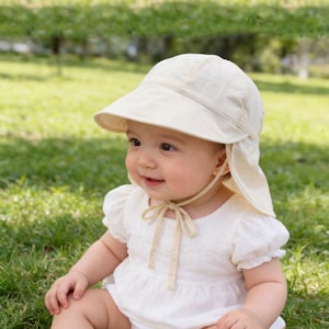 May include: A baby wearing a cream-coloured sun hat with a wide brim and neck flaps. The baby is also wearing a white, short-sleeved romper with lace detailing. The background is a blurred green grassy area.