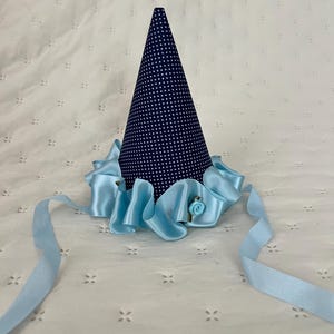 May include: A conical party hat featuring a dark blue fabric with a white polka dot pattern. The base of the cone is adorned with a ruffled light blue satin ribbon trim and a small light blue fabric rose. Two long light blue satin ribbons extend from the base.
