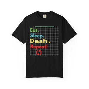 Puede incluir: Camiseta negra con la frase "Eat. Sleep. Dash. Repeat!" en un diseño de cuadrícula. El texto es en verde, azul, amarillo y rojo. Una línea verde está encima de "Eat", y un símbolo de reciclaje rojo está debajo de "Repeat!"