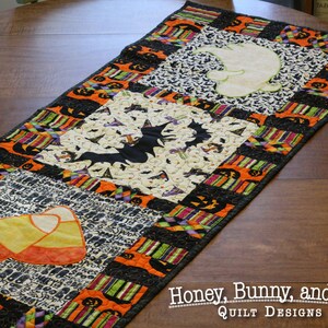 May include: A black and orange Halloween table runner with a candy corn, bat, and ghost design. The runner is made of fabric and has a quilted texture. "Honey, Bunny, and Doll Quilt Designs" is printed on the table.