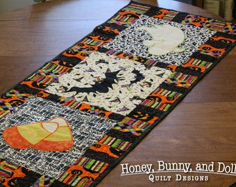 May include: A black and orange Halloween table runner with a candy corn, bat, and ghost design. The runner is made of fabric and has a quilted texture. "Honey, Bunny, and Doll Quilt Designs" is printed on the table.