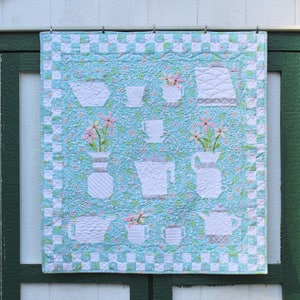 May include: A blue and white quilted wall hanging with a checkered border. The design features a variety of white teapots, cups, and vases with pink flowers.