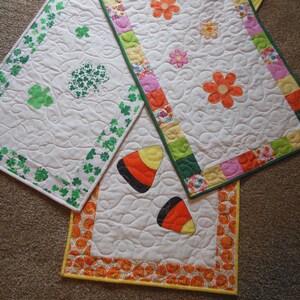 May include: Three quilted table runners with different designs. The first runner has a green shamrock pattern, the second has a colorful floral pattern, and the third has a yellow and orange pumpkin pattern.