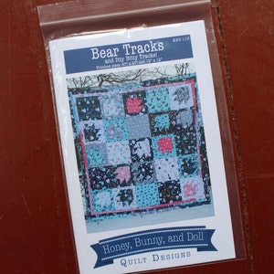 May include: A quilt pattern titled "Bear Tracks and Itty Bitty Tracks!" is displayed in a clear plastic bag. The quilt design features bear paw prints in pink, blue, and gray squares. Finished sizes are 47" x 47" and 12" x 12". The brand is Honey, Bunny, and Doll.