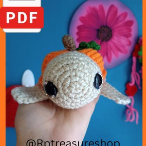 May include: A small, handmade crochet turtle toy featuring a cream-colored head with large, shiny black plastic eyes, an orange shell, and a small brown stem on top. The text @Rptreasureshop is visible at the bottom of the image.