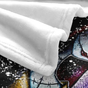 Peut inclure: Gros plan sur une couverture blanche et moelleuse avec une illustration d&eacute;taill&eacute;e. Le bord de la couverture est visible, r&eacute;v&eacute;lant une surface douce et textur&eacute;e. L'illustration pr&eacute;sente un personnage avec des traits cousus et un fond sombre.