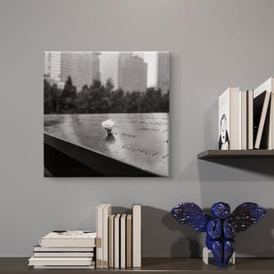 May include: Black and white photograph of a memorial with a single white flower. The memorial is inscribed with names and set against a backdrop of city buildings. The image is displayed on a canvas print.
