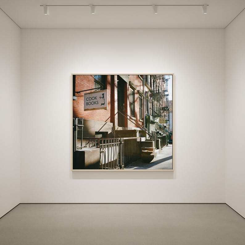 May include: A framed photograph of a New York City street scene. The image features a brick building with a sign that reads "COOK BOOKS" and a stylized image of a chef. The scene is bathed in sunlight, with shadows cast across the steps and sidewalk.