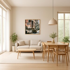 May include: A living room with a neutral color palette. A framed picture of a street scene hangs above a beige sofa. A wooden coffee table and dining set are in the foreground. A large window and potted plants add natural light and greenery.