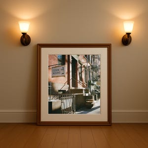 May include: A framed print of a New York City street scene, featuring a brick building with a sign that reads "COOK BOOKS." The print is displayed on a wooden floor, with two wall sconces on either side.