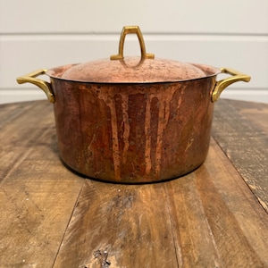 May include: A copper cooking pot with a lid. The pot has a round body with a tarnished, aged appearance. It features gold-colored handles and a matching handle on the lid. The pot sits on a wooden surface.