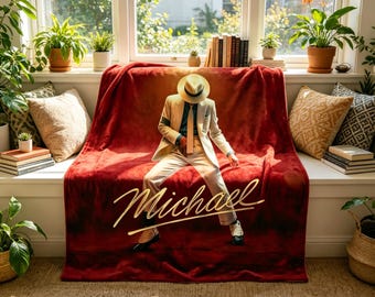 May include: A red plush throw blanket featuring a gold script of the name "Michael" and an image of a person in a cream-colored suit, hat, and tie. The blanket is draped over a red bench in front of a window.