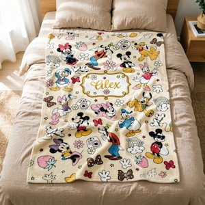 Könnte beinhalten: Eine cremefarbene Fleecedecke mit einem wiederkehrenden Muster aus Disney-Figuren wie Mickey Maus, Minnie Maus, Donald Duck, Daisy Duck, Goofy und Pluto, durchsetzt mit Erdbeeren, Regenbögen und Kameras, sowie dem Namen Alex in goldener Schrift.