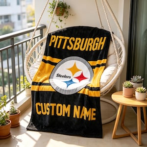 May include: A black and yellow fleece blanket featuring the Pittsburgh Steelers logo in the center, the word PITTSBURGH at the top, and the text CUSTOM NAME at the bottom. The blanket has horizontal yellow stripes across the black fabric.