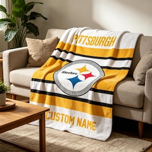 May include: A soft fleece throw blanket featuring the Pittsburgh Steelers logo in the center, with horizontal yellow, white, and black stripes. The word PITTSBURGH is printed in yellow at the top, and the text CUSTOM NAME appears at the bottom.