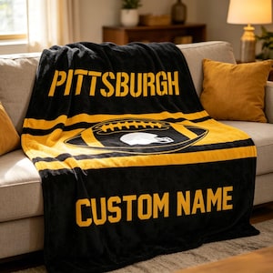 May include: A black and yellow fleece throw blanket featuring the word PITTSBURGH in bold yellow letters at the top, a central graphic of a football with a white helmet silhouette, and the text CUSTOM NAME in yellow letters at the bottom.