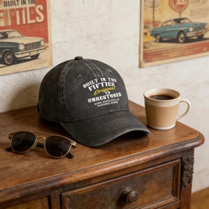 May include: A black baseball cap with the text "BUILT IN THE FIFTIES Original & UNRESTORED" in white and yellow. The cap is on a wooden table with sunglasses and a coffee mug. Vintage theme.