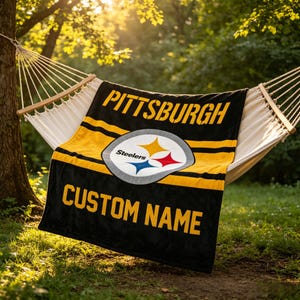 May include: A black and yellow fleece blanket featuring the Pittsburgh Steelers logo in the center, the word PITTSBURGH in yellow text at the top, and the words CUSTOM NAME in yellow text at the bottom, draped over a white rope hammock.