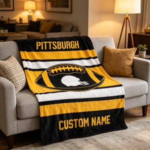 May include: A black, yellow, and white throw blanket featuring the text PITTSBURGH at the top and CUSTOM NAME at the bottom. The center displays a large graphic of a football with a white helmet silhouette inside, set against horizontal stripes.