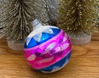 May include: A round glass Christmas ornament featuring horizontal bands of bright blue and vibrant pink, accented with silver glitter star patterns. The ornament has a silver metal cap and is positioned in front of several bottle brush trees.